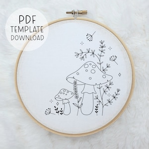 May include: A black and white line drawing of two mushrooms with stems and caps, surrounded by small flowers and leaves. The image is on a white background and is in a wooden embroidery hoop.