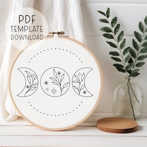 Puede incluir: Un aro de bordado blanco con un dibujo de línea negra de tres lunas crecientes con ramas y estrellas dentro de cada luna. El aro está sobre una superficie de madera blanca.