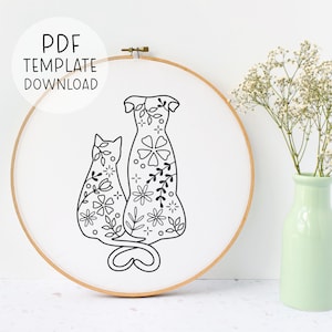 Könnte beinhalten: Schwarzweißes Linienkunst-Stickmuster mit einem Katz und einem Hund mit floralen Details. Das Muster ist auf einem weißen Stoff in einem hölzernen Stickrahmen dargestellt.