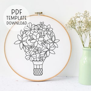 Puede incluir: Diseño de arte de línea en blanco y negro de un globo aerostático lleno de flores. El diseño está en un lienzo blanco en un aro de bordado de madera.