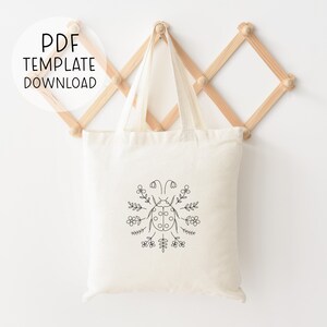 Puede incluir: Una bolsa de lona blanca con un dibujo de l&iacute;nea negra de una mariquita rodeada de flores. El texto "PDF TEMPLATE DOWNLOAD" est&aacute; en un c&iacute;rculo en la parte superior de la imagen.