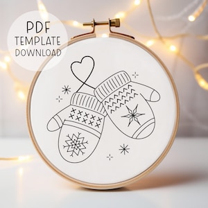 May include: A round embroidery hoop with a white fabric featuring a black and white design of two mittens connected by a heart-shaped line. The text "PDF TEMPLATE DOWNLOAD" is at the top. The design includes snowflakes and star details.