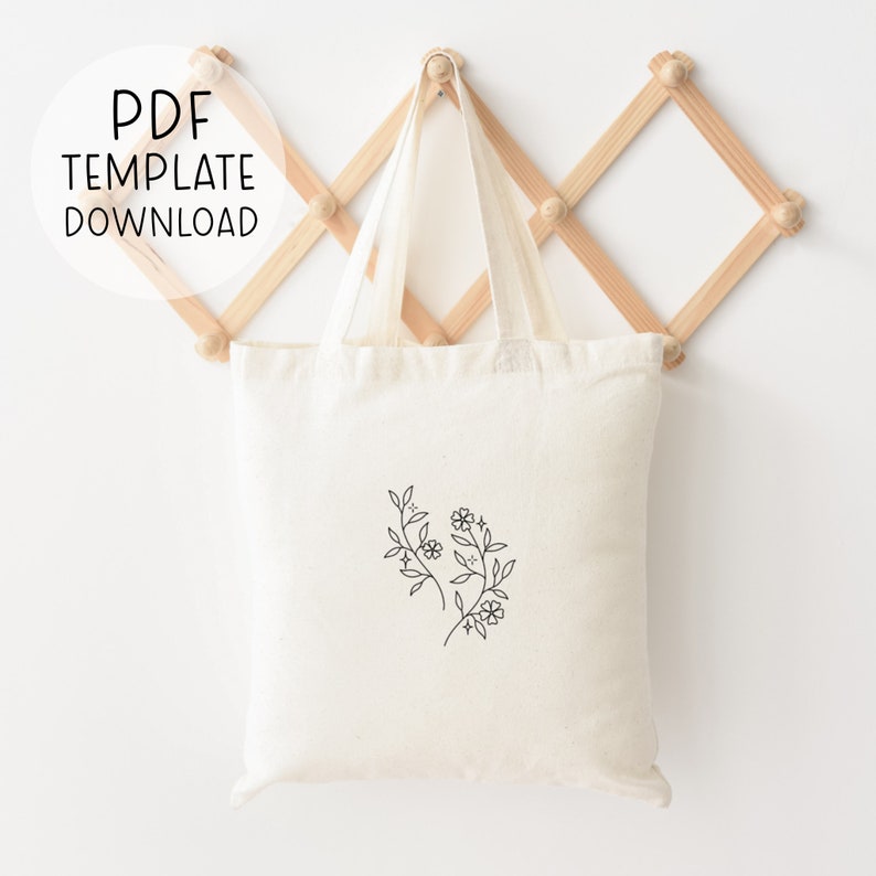 Pode incluir: Uma bolsa tote de lona branca com um desenho simples de dois ramos florais com pequenas flores na frente. A bolsa est&aacute; pendurada em um rack de madeira.