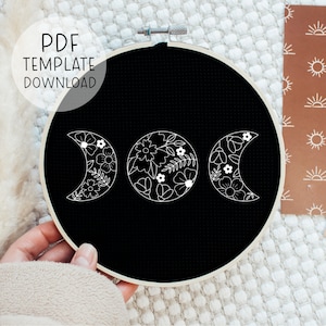 Puede incluir: Un aro de bordado negro con un diseño de fase lunar floral blanco. El diseño presenta tres lunas, cada una con una fase diferente, y cada luna está llena de flores y hojas.