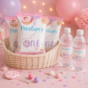May include: Three snack bags and two water bottles with custom labels featuring the text Penelope's sweet ONE, decorated with illustrations of donuts, cupcakes, and pink icing drips. The items are arranged in a light-colored woven basket.