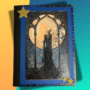 May include: A decorative journal with a blue border and a fantasy-themed image. The image features a woman in a long black dress standing before a large arched window with a full moon. Gold glitter star embellishments adorn the cover.