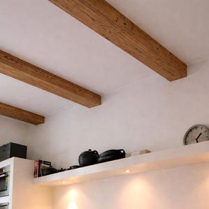 Può includere: Tre travi a soffitto rettangolari in legno con finitura a venature naturali installate contro un soffitto bianco. Sotto le travi, una parete bianca presenta una mensola integrata che ospita utensili neri e un orologio da parete rotondo.