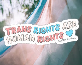 May include: A sticker with the text "Trans Rights Are Human Rights" in pink, blue, and white lettering with a blue heart. The sticker is on a background of a blue, pink, and white fabric.