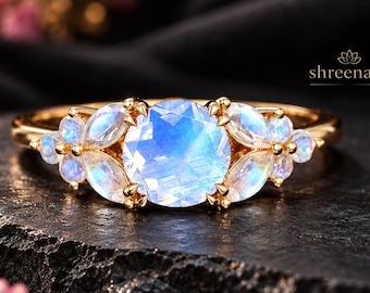 May include: A gold ring featuring a large round faceted moonstone center stone flanked by smaller marquise and round moonstones. The text shreenath is visible in the upper right corner. The ring has a polished gold band and prong settings.