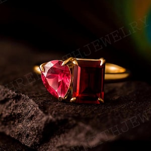 May include: A gold ring featuring two red gemstones: a heart-shaped stone and a rectangular stone. The ring is set against a dark, textured background, with a blurred peacock feather in the background.