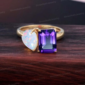 May include: A gold ring featuring a heart-shaped opal and a square-shaped amethyst gemstone. The opal is white with iridescent colours, and the amethyst is a deep purple.