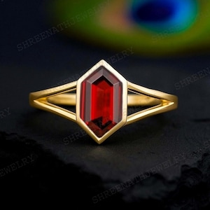 May include: A gold ring with a geometric design, featuring a large, rectangular, deep red gemstone. The ring's band splits into two delicate strands on either side of the gemstone, creating an open, airy design. The ring is set against a dark, textured background.