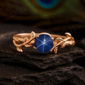 May include: A rose gold ring featuring a round, deep blue star sapphire gemstone. The ring's band is adorned with delicate leaf and vine detailing, creating a nature-inspired design. The gemstone has a prominent six-ray star pattern.