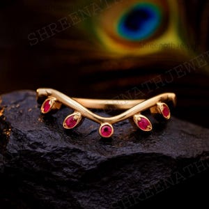 May include: A gold-colored ring with a curved design, featuring five red gemstones. The ring is set on a black surface, with a blurred background. The gemstones are oval-shaped, and the ring appears to be made of metal.
