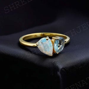 May include: A gold ring with two teardrop-shaped gemstones. The larger gemstone is a clear, iridescent moonstone. The smaller gemstone is a light blue aquamarine.