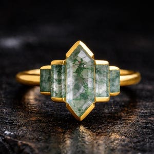 May include: A gold ring featuring a central, clear, emerald-cut gemstone with green inclusions, flanked by two smaller rectangular green stones on each side. The ring has a gold band and setting, set against a dark, textured background.