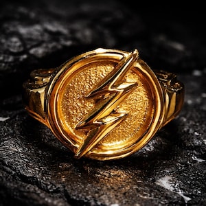 May include: A gold-colored ring featuring the Flash symbol, a lightning bolt within a circle. The ring has a textured surface and a polished finish, set against a dark, textured background. A piece of jewelry.