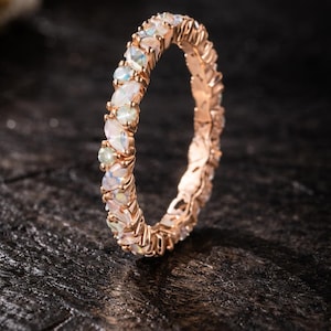 May include: Rose gold ring with a continuous band of oval-shaped, iridescent gemstones. The ring's setting is a warm, metallic hue, and the gemstones display a range of colors. The ring is set against a dark, textured background.