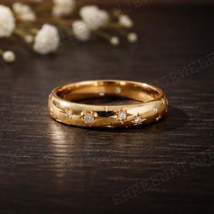 Puede incluir: Un anillo dorado con diseños en forma de estrella, cada uno con un pequeño diamante brillante. El anillo se muestra sobre una superficie de madera oscura, con flores blancas en el fondo. La banda del anillo mide aproximadamente 0,5 cm de ancho.
