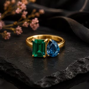 Könnte beinhalten: Ein Goldring mit zwei markanten Edelsteinen: einem rechteckigen, smaragdgeschliffenen grünen Stein und einem tropfenförmigen blauen Stein. Die Steine sind nebeneinander auf einer polierten Goldschiene gefasst und bilden ein auffälliges Design.