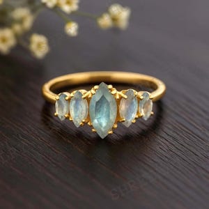 May include: A gold ring featuring a central marquise-cut, light blue gemstone flanked by four smaller oval-cut gemstones. The ring's band is a simple, polished gold. The gemstones have a subtle, iridescent quality. The ring is set against a dark wooden surface.