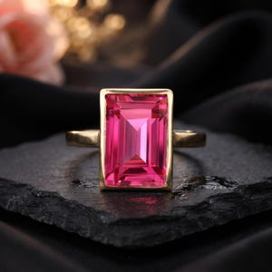 May include: A gold ring featuring a large, rectangular, emerald-cut vibrant pink gemstone set in a smooth, polished gold bezel. The ring band is simple and metallic, showcasing a clean design that highlights the faceted pink stone.