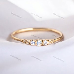 May include: A delicate gold band ring with five small, clear gemstones set in a curved line. The ring is on a white fabric background.