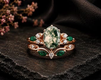 Könnte beinhalten: Ein Ring-Set aus Roségold mit einem zentralen ovalen Moosachat, akzentuiert durch kleine runde klare Steine und grüne Steine im Marquise-Schliff. Die Ringschiene hat ein geschwungenes Design mit weiteren klaren und grünen Steinen.