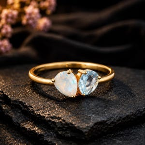 May include: A gold ring featuring two pear-shaped gemstones set side-by-side. One stone is an iridescent white opal, and the other is a faceted light blue aquamarine. The band is a smooth, polished gold metal, creating a delicate and refined look.
