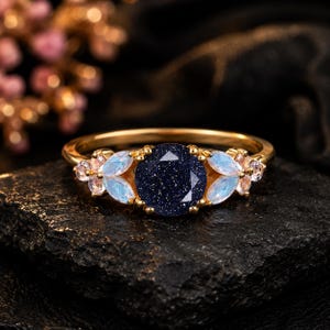 Op de afbeelding: Een gouden ring met een ronde, donkerblauwe, glinsterende zandsteen in het midden. De centrale steen wordt aan weerszijden geflankeerd door twee marquise-geslepen, iriserende, lichtblauwe stenen en een kleine, heldere, ronde accentsteen.