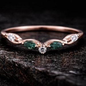 Puede incluir: Anillo de oro rosa con un diseño curvo. El anillo presenta tres piedras verdes con corte marquesa y dos piedras blancas redondas. Una sola piedra blanca redonda está centrada en el ápice del anillo. El anillo está sobre una superficie oscura y texturizada.