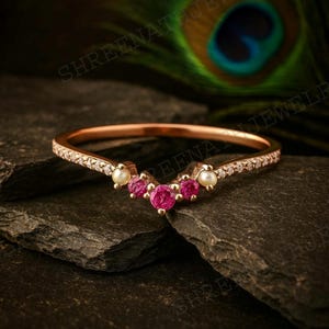 May include: A rose gold ring with a curved design. The ring features small, round, pink gemstones and pearls, with a band encrusted with small clear stones. The ring is set against a dark, textured background.