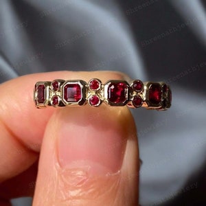 Könnte beinhalten: Ein Goldring mit einer Reihe von roten quadratischen Edelsteinen und kleineren runden roten Edelsteinen.