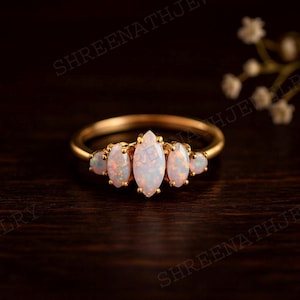 May include: A gold ring featuring five opal gemstones. The central opal is marquise-cut, flanked by two oval opals on each side. The opals display a milky white color with flashes of orange and pink.