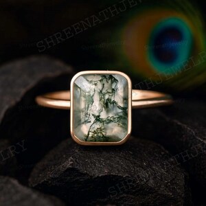 Pode incluir: Um anel de ouro com uma pedra preciosa de ágata musgo retangular. A pedra preciosa tem uma aparência translúcida com inclusões verdes semelhantes a musgo. O anel está sobre uma superfície escura e texturizada.