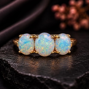 Può includere: Un anello in oro con tre opali di forma ovale disposti in fila. Ogni opale presenta una base bianco latte con riflessi iridescenti blu, verdi e rosa. Le pietre sono fissate da piccole griffe in oro su una fascia in oro lucido.