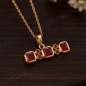Peut inclure: Un collier en or avec un pendentif orné de trois pierres précieuses rouges carrées et de petites pierres précieuses rouges rondes. Le pendentif est suspendu à une fine chaîne en or, sur un fond en bois sombre.