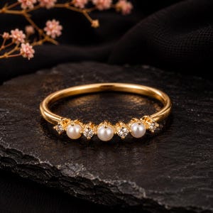 Op de afbeelding: Een gouden ring met een fijne band, bezet met drie kleine witte parels afgewisseld met vier heldere, fonkelende ronde stenen. Het sieraad heeft een gepolijste metalen afwerking en een symmetrische rangschikking van de edelstenen aan de voorzijde.
