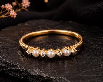 Puede incluir: Un anillo de oro con una banda delicada engastada con tres pequeñas perlas blancas que se alternan con cuatro piedras redondas transparentes y brillantes. La pieza de joyería muestra un acabado metálico pulido y una disposición simétrica.