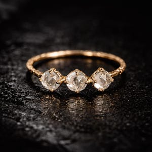 May include: A gold ring featuring three clear, round gemstones. The ring band has a textured pattern. The gemstones are set in a gold setting, and the ring is shown on a dark, textured surface.
