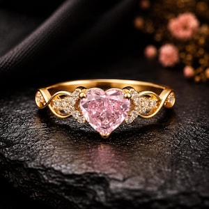 May include: A gold ring featuring a central heart-shaped pink gemstone. The band is crafted with an intricate twisted design and is accented with small, clear pavé-set stones on both sides of the heart, creating a detailed and elegant appearance.