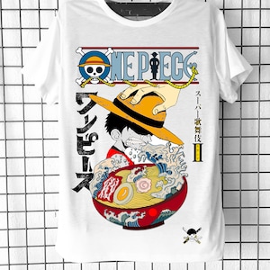 Peut inclure: T-shirt blanc avec un imprimé graphique d'un personnage de dessin animé de l'anime One Piece. Le personnage mange un bol de ramen avec un motif de vague. Le texte "ONE PIECE" est imprimé en rouge et noir au-dessus du personnage. Le texte "ワンピース" est imprimé en noir sur le côté gauche du personnage.