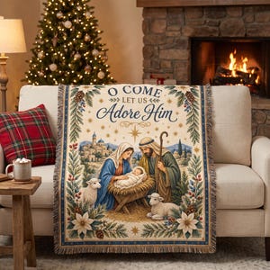 May include: A woven throw blanket featuring a nativity scene with Mary, Joseph, and the baby Jesus in a manger. The text O COME LET US ADORE HIM is printed at the top. The design is framed by pine branches, pinecones, and white poinsettias.