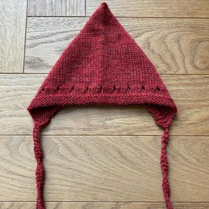 Könnte beinhalten: Eine handgestrickte, dreieckige Mütze in tiefroter Farbe. Die Mütze hat Ohrenklappen mit gedrehten Bändern und eine dekorative Reihe kleiner Löcher am Rand. Die Mütze ist aus strukturiertem Garn gefertigt.
