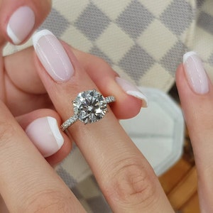 Pu&ograve; includere: Primo piano di un anello di fidanzamento con diamante, con una pietra centrale a taglio brillante rotondo e una fascia pav&eacute; di diamanti. L'anello &egrave; su un dito con una manicure bianca.