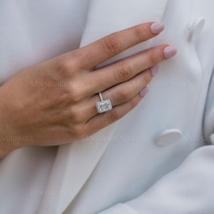 May include: A close-up of a hand wearing a silver ring with a large, rectangular, clear gemstone. The ring has a halo of smaller gemstones around the center stone.