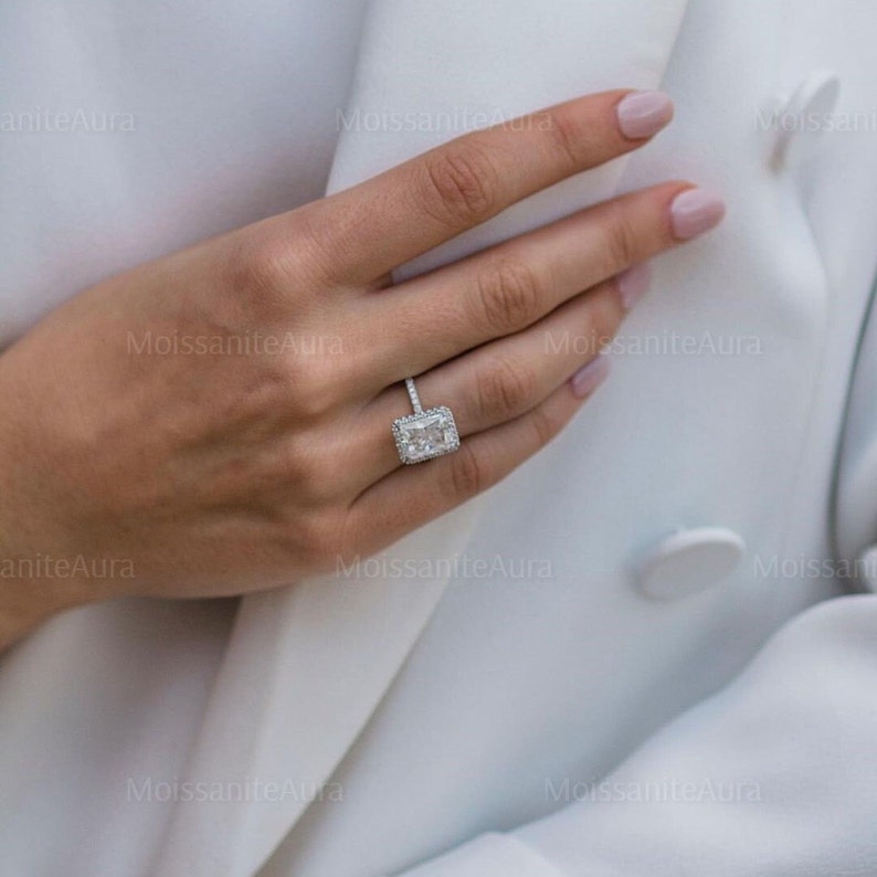 May include: A close-up of a hand wearing a silver ring with a large, rectangular, clear gemstone. The ring has a halo of smaller gemstones around the center stone.
