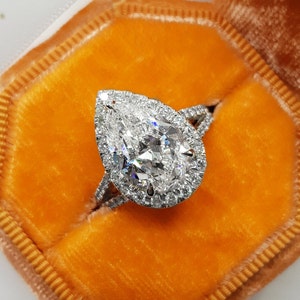 Op de afbeelding: Een peervormige diamanten verlovingsring met een halo-setting en een dubbele band. De ring is gezet in een witgouden band en wordt gepresenteerd in een oranje fluwelen ringdoos.