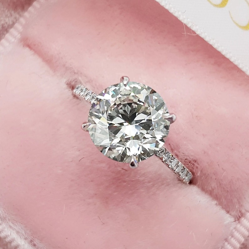 Pu&ograve; includere: Un anello di fidanzamento con diamante rotondo e chiaro con una fascia d'argento e una fila di diamanti pi&ugrave; piccoli attorno alla pietra centrale.