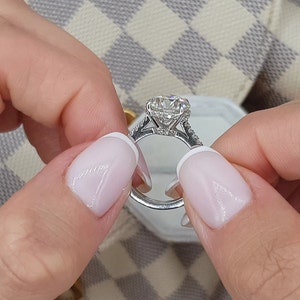 Pu&ograve; includere: Primo piano di un anello di fidanzamento in argento con un grande diamante rotondo e chiaro al centro. L'anello ha una fascia di diamanti pav&eacute; e una montatura a halo.